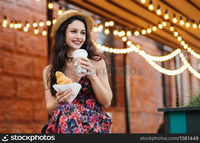 Portrait of restful woman with dark hair, shining eyes and well-shaped lips wearing hat and dress, having dinner, eatting croissant and drinking coffee looking aside with smile, having good mood