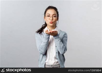Portrait of romantic and passionate, pretty asian girlfriend in glasses send, blowing air kiss to camera with hands near folded lips, express symapthy, romance and flirt, stand grey background.. Portrait of romantic and passionate, pretty asian girlfriend in glasses send, blowing air kiss to camera with hands near folded lips, express symapthy, romance and flirt, stand grey background