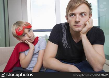 Portrait of sad boy dressed in superhero costume sitting with father on sofa bed at home