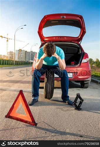 Portrait of sad man sitting on spare wheel near broken car