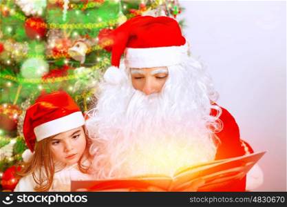 Portrait of Santa Claus with cute granddaughter sitting near Christmas tree, studio shot, reading magic book, celebrating winter holidays