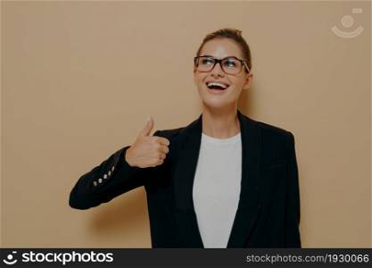 Portrait of satisfied female in eyeglasses smiling while showing thumb up with hand and looking upside with positive expression, demonstrating agreement while posing isolated over beige background. Portrait of satisfied female in eyeglasses smiling while showing thumb up with hand