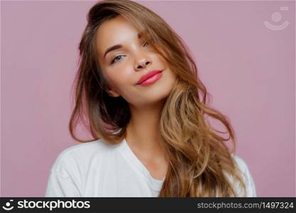 Portrait of satisfied relaxed young female model tilts head, has makeup, fair hair, dressed in white clothes, poses against purple background, has well cared complexion. People, beauty, face care