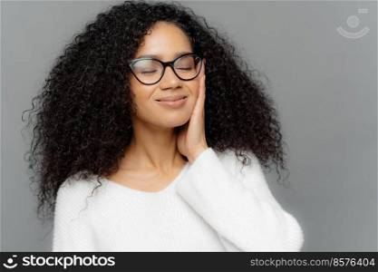 Portrait of satsfied dark skinned woman with curly hair, has eyes closed, touches cheek with hands, imagines something pleasant, wears optical glasses and white jumper, poses indoor. Close up portrait