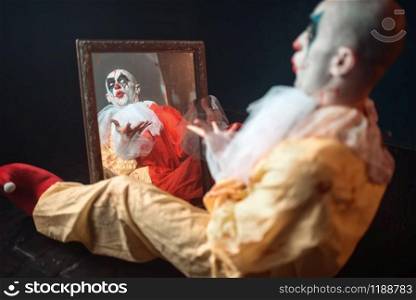 Portrait of scary bloody clown with crazy eyes sitting at the mirror, nightmare. Man with makeup in carnival costume, mad maniac. Bloody clown with crazy eyes sitting at the mirror