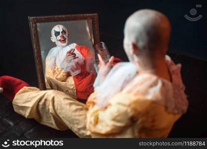 Portrait of scary bloody clown with crazy eyes sitting at the mirror, nightmare. Man with makeup in carnival costume, mad maniac