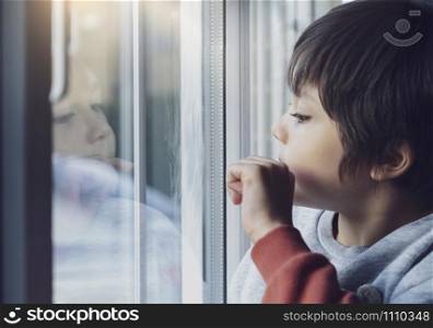 Portrait of school boy sitting next to window and using his finger drawing or writing on window glass, Side view of child hand drawing on glass, Toddler boy looking at him self through window.