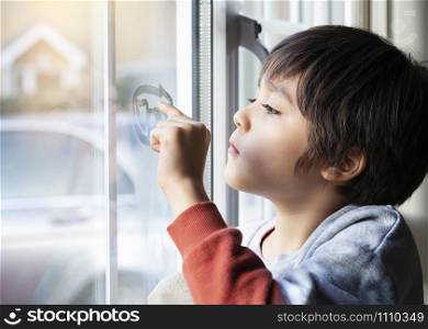Portrait of school boy sitting next to window and using his finger drawing two hearts on window, Side view of child hand writing on glass, Toddler boy looking at him self through window.