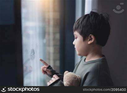 Portrait of school boy standing next to window and using his finger drawing or writing on window glass, Side view of child hand drawing on glass, Toddler boy looking at him self through window.