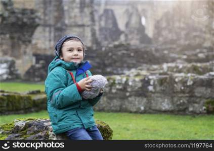 Portrait of School kid taking teddy bear explore with his learning history, Happy child boy wearing warm cloths holding his soft toy sitting on old brick wall with blurry ruins of old abbey
