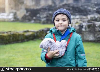 Portrait of School kid taking teddy bear explore with his learning history, Happy child boy wearing warm cloths holding his soft toy standing alone with blurry ruins of old abbey background