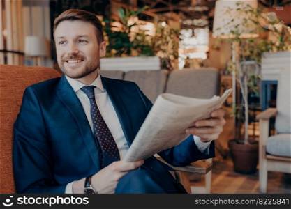 Portrait of seated successful young entrepreneur in modern formal clothes while reading section of financial chronicle in newspaper, waiting for ordered drink on cozy sofa in cafe in morning. Portrait of successful young entrepreneur in formal suit sitting in cafe