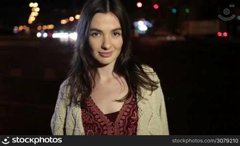 Portrait of seductive brunette woman flirting with camera and looking at someone temptingly over night city streetlights and traffic background. Sexy beautiful girl with enigmatic look posing on street at night . Slow motion. Stabilized shot.