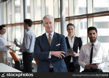 portrait of senior businessman as leader at modern bright office interior, young people group in background as team