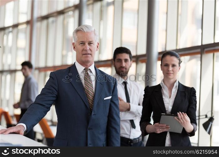 portrait of senior businessman as leader at modern bright office interior, young people group in background as team