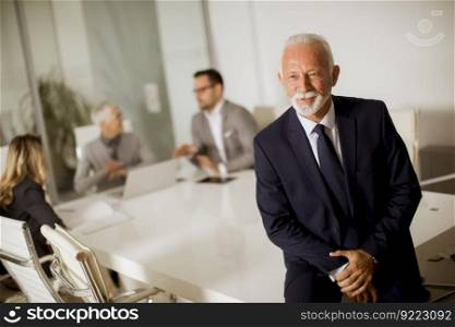 Portrait of senior businessman standing in office while other business people having meeting