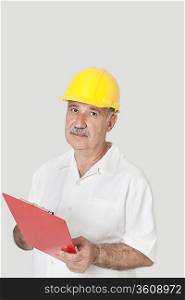 Portrait of senior constructor standing with clipboard over gray background