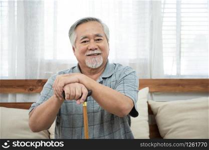 Portrait of senior old elderly asian man sit on coach hand hold help walking stick sit on sofa in house look at camera with happiness and healthy lifestyle.