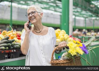 Portrait of senior woman buying on market,  holding basket with flowers and using mobile phone