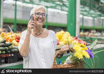 Portrait of senior woman buying on market,  holding basket with flowers and using mobile phone