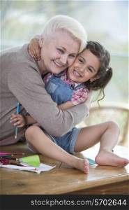 Portrait of senior woman hugging granddaughter sitting on table at home