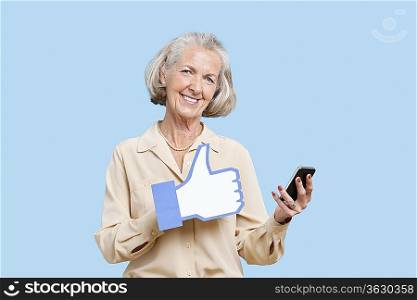 Portrait of senior woman with cell phone holding fake like button against blue background