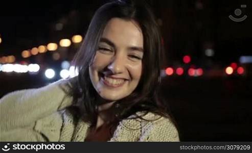 Portrait of sensual brunette female playing with camera on night city street and smiling. Seductive young woman with amazing long brown hair posing on city street in colorful bokeh of streetlights background. Slow motion. Steadicam stabilized shot.