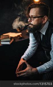 Portrait of serious bearded man in glasses smoking pipe, table with books on background. Writer, journalist, literature author, blogger or poet concept. Serious bearded man in glasses smoking pipe