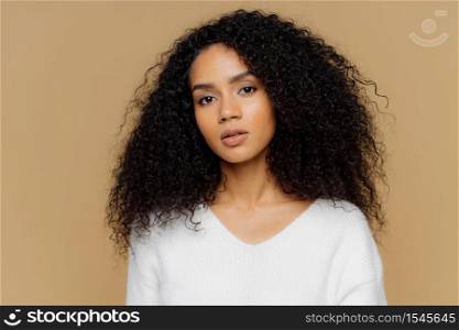 Portrait of serious beautiful dark skinned female with frizzy black hair, has minimal makeup, looks calmly at camera, wears white jumper, stands against brown background, being deep in thoughts.