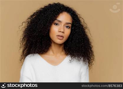 Portrait of serious beautiful dark skinned female with frizzy black hair, has minimal makeup, looks calmly at camera, wears white jumper, stands against brown background, being deep in thoughts.