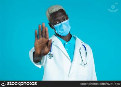 Portrait of serious black doctor in professional medical white coat and mask showing rejecting gesture by stop palm sign. Doc man isolated on blue background. Portrait of serious black doctor in professional medical white coat and mask showing rejecting gesture by stop palm sign. Doc man isolated on blue background.