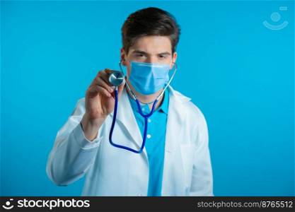 Portrait of serious doctor in professional medical coat and mask using stethoscope isolated on blue studio background. Man points his instrument at camera as if he is listening to ill patient.. Portrait of serious doctor in professional medical coat and mask using stethoscope isolated on blue studio background. Man points his instrument at camera as if he is listening to ill patient
