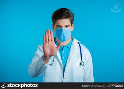 Portrait of serious doctor in professional medical white coat showing rejecting gesture by stop palm sign. Doc man isolated on blue background. High quality photo. Portrait of serious doctor in professional medical white coat and mask showing rejecting gesture by stop palm sign. Doc man isolated on blue background.