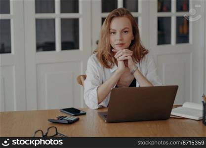 Portrait of serious female professional manager sitting at laptop at office desk, looking at camera. Confident pensive young businesswoman using computer for remote job posing on workplace. . Portrait of confident businesswoman sitting at laptop at office desk, looking at camera on workplace