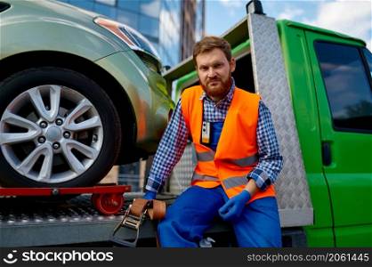 Portrait of serious male tow truck operator holding fixing belt sitting on evacuator platform. Portrait of serious male tow truck operator