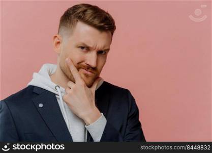 Portrait of serious man holds chin looks displeased at camera being deep in thoughts wears stylish clothes tries to make decision isolated over pink background with blank space for your promo