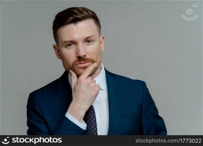 Portrait of serious professional businessman ceo executive in suit touching his chin with stubble and thinking about business startup while standing against grey studio background, close up portrait. Confident handsome male entrepre thinking about business