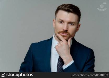 Portrait of serious professional businessman ceo executive in suit touching his chin with stubble and thinking about business startup while standing against grey studio background, close up portrait. Confident handsome male entrepre thinking about business