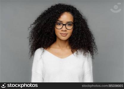 Portrait of serious woman with dark skin, Afro bushy hair, wears big transparent glasses and white soft sweater, looks directly at camera, isolated over grey background. People and ethnicity concept