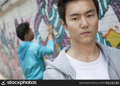 Portrait of serious young man with a cool attitude looking at the camera, young man spray painting in the background