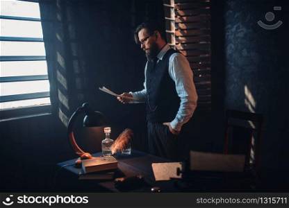 Portrait of seriuos bearded man in glasses reads handwritten text. Desk with retro lighting lamp on background. Writer, journalist, literature author, blogger or poet concept. Bearded man in glasses reads handwritten text