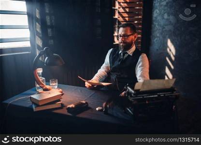 Portrait of seriuos bearded man in glasses reads handwritten text. Desk with retro lighting lamp on background. Writer, journalist, literature author, blogger or poet concept. Bearded man in glasses reads handwritten text