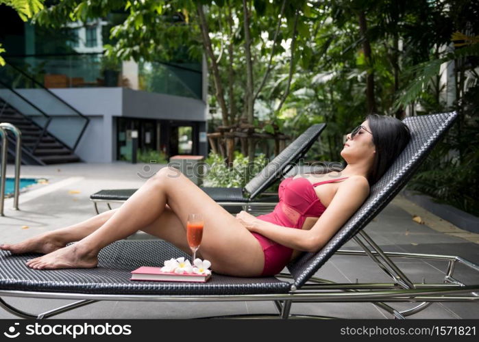 Portrait of sexy Asian beautiful woman in red swimsuit and sunglasses sleep or nap on bed chair to get suntan near swimming pool. Female relax in luxury hotel for summer holiday or vacation.