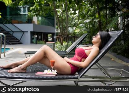 Portrait of sexy Asian beautiful woman in red swimsuit and sunglasses sleep or nap on bed chair to get suntan near swimming pool. Female relax in luxury hotel for summer holiday or vacation.