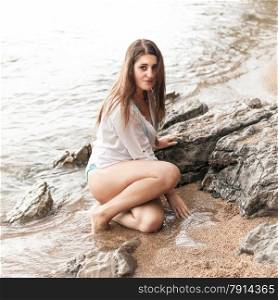 Portrait of sexy brunette woman in wet shirt sitting next to big rock at sea