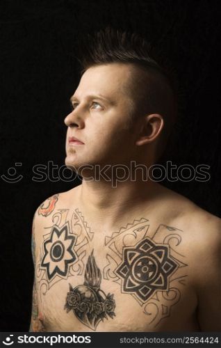 Portrait of shirtless Caucasian man with tattoos and mohawk against black background.