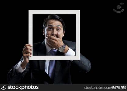 Portrait of shocked businessman covering mouth while holding frame against black background