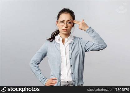 Portrait of sick and tired asian woman hear something stupid, shoot herself with finger gun as a reaction to annoying bothering conversation, staring skeptical camera, standing grey background.. Portrait of sick and tired asian woman hear something stupid, shoot herself with finger gun as a reaction to annoying bothering conversation, staring skeptical camera, standing grey background