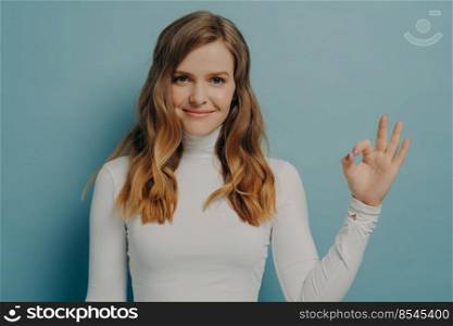 Portrait of sitisfied young europian woman showing okay gesture with fingers isolated over pastel blue stuio wall background, female demonstrating approvement. Positive body language concept. Cheerful young european woman showing okay gesture isolated over pastel blue studio wall