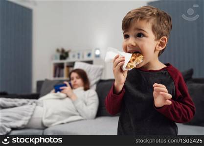 Portrait of small boy caucasian little child kid having dinner sandwich in napkin or pastry holding in front of opened mouth eating in the evening his mother is lying on bed in back plan defocused